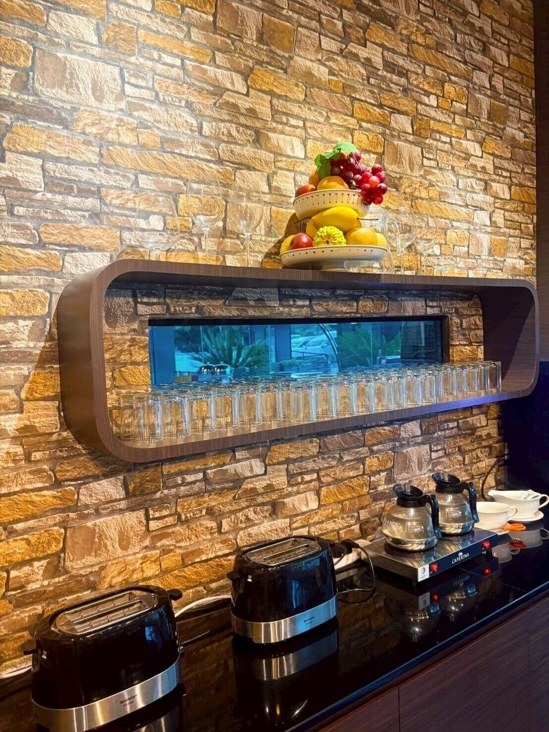 A kitchen with a stone wall, a fruit display, a glass shelf with glasses, a toaster, and a coffee maker.
