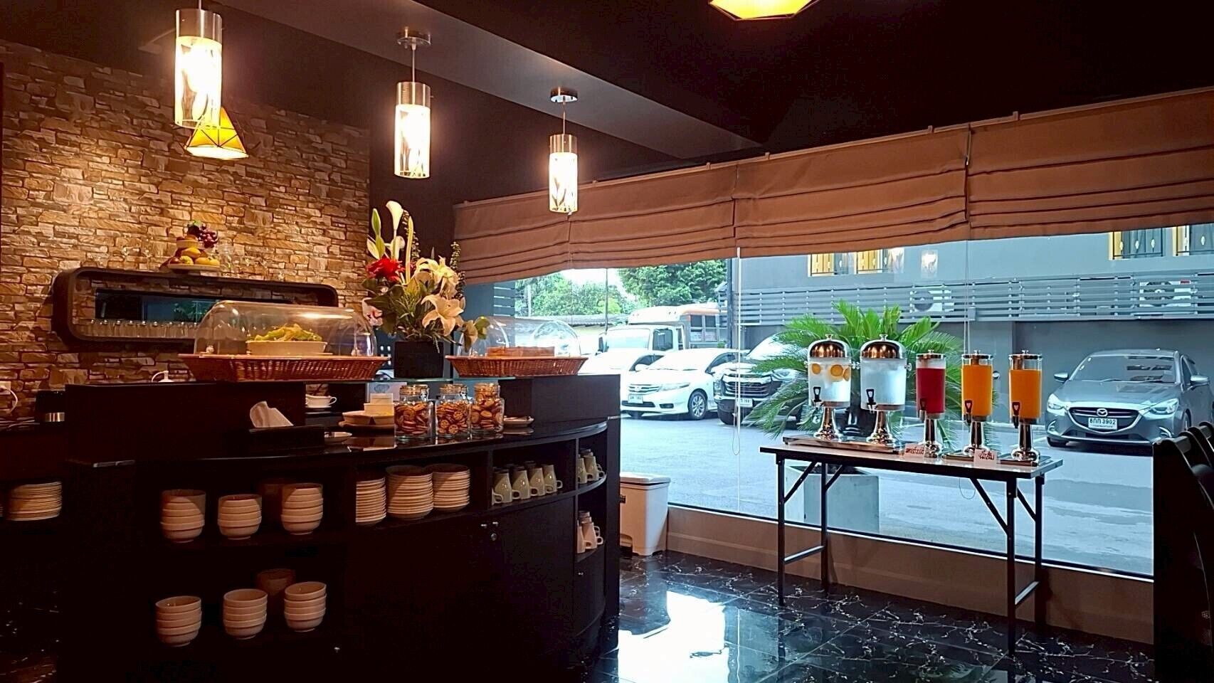 A breakfast bar with a stone wall, a window view of parked cars, and a table with juice dispensers.