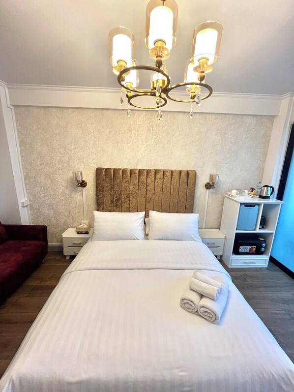 Una habitación de hotel con una cama grande, cabecera de madera, mesitas de noche con lámparas, un sillón rojo y un minibar.