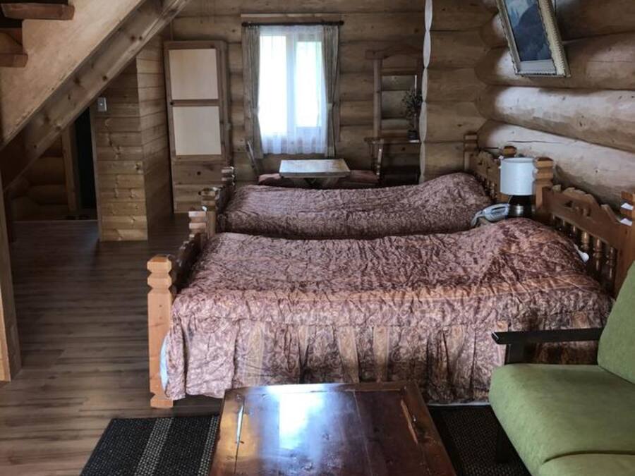 Ein Schlafzimmer in einer Blockhütte mit einem großen Bett, einem grünen Sessel, einem Holztisch und einem Fenster mit Vorhängen.