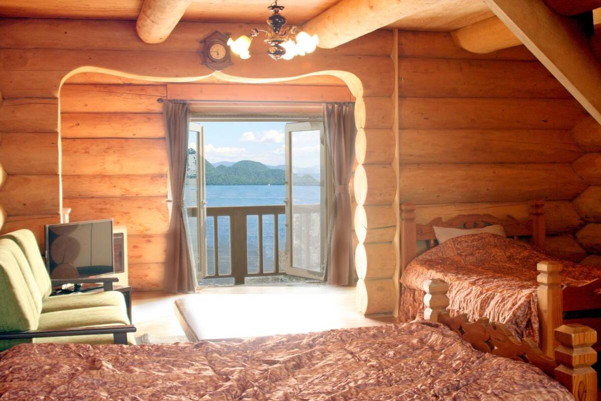 A log cabin room with a bed, a chair, a television, and a balcony with a view of the water.