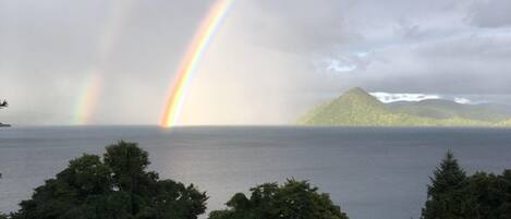 A view of a coastal landscape with a rainbow over the sea, a mountain in the background, and a house in the foreground.