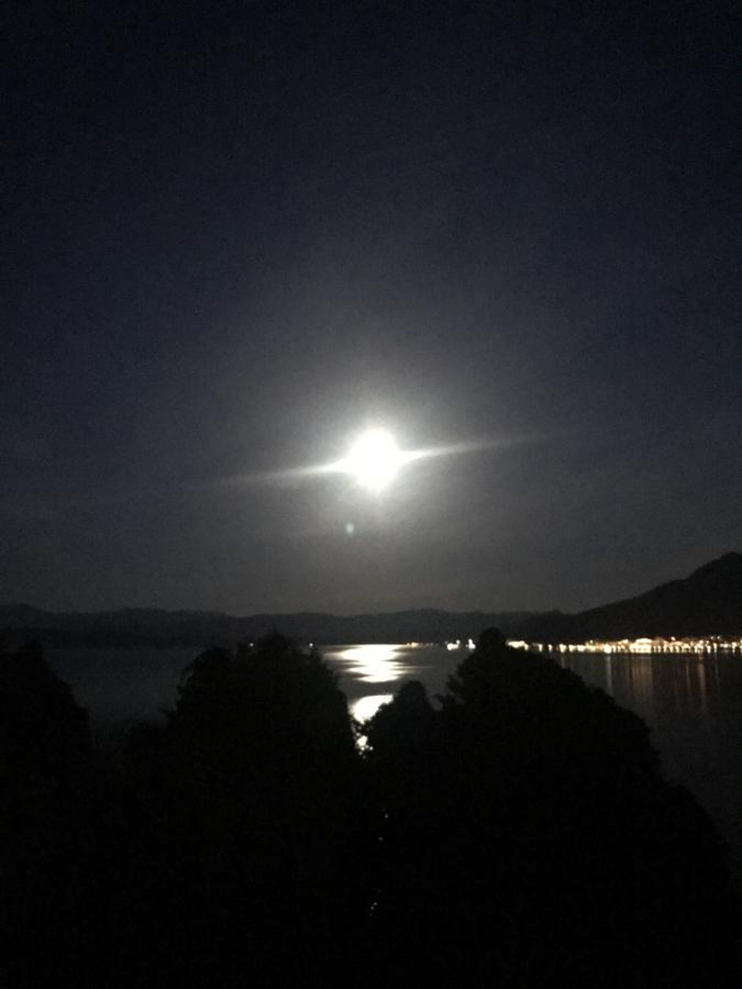 A night scene with a bright moon or light source in the sky, a body of water, and distant lights along a shoreline.