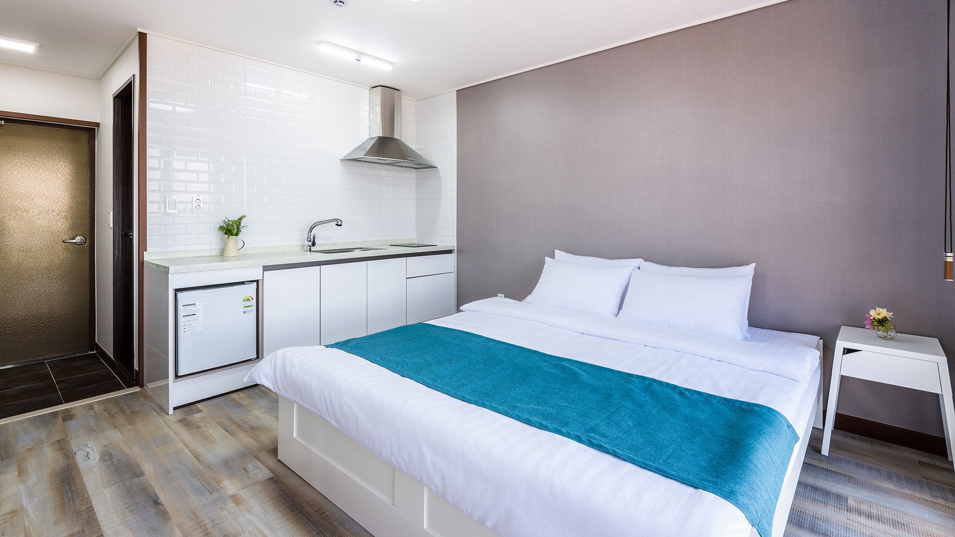 A modern hotel room with a bed, a small bedside table, a nightstand, a built-in kitchen with a sink, and a shower area.