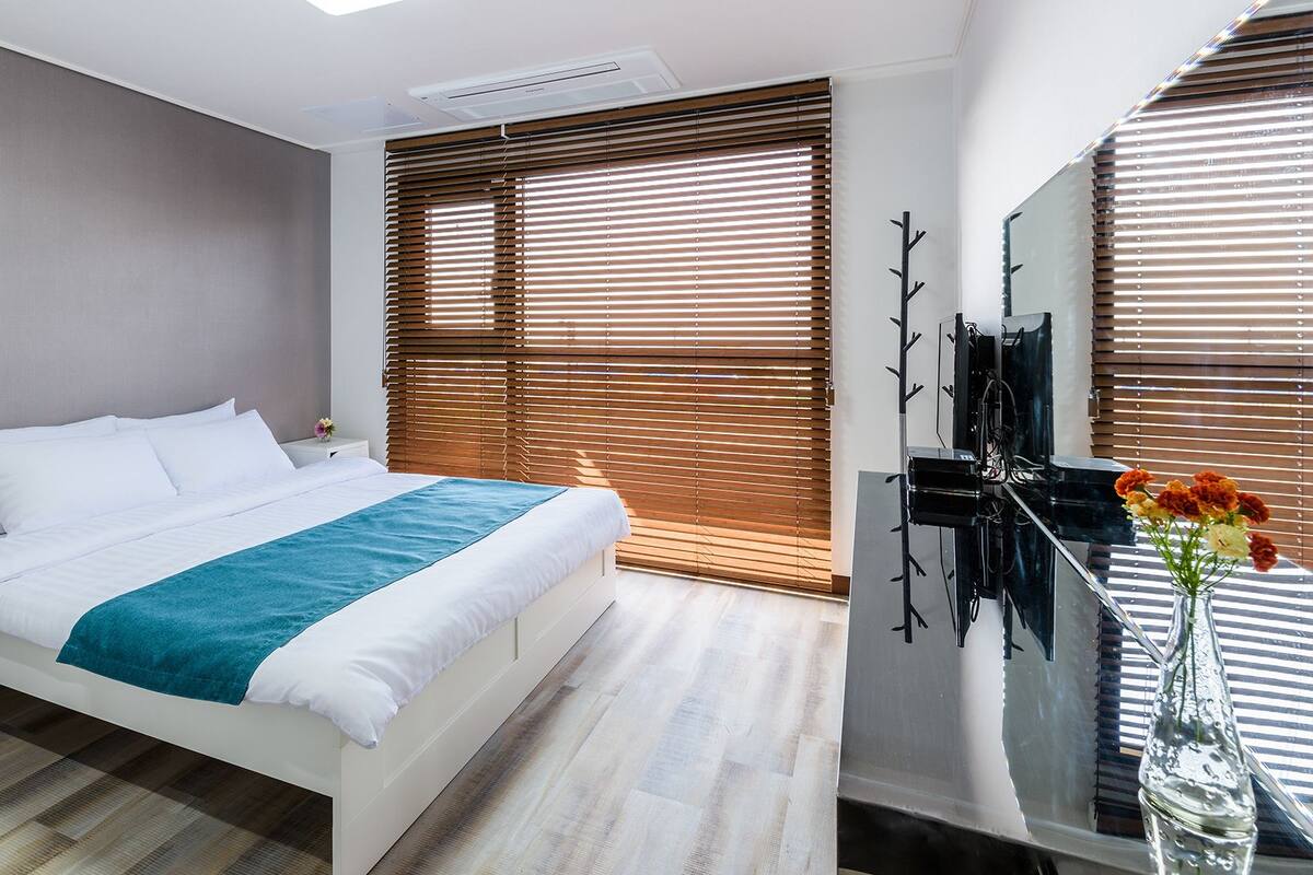 A modern hotel room with a large bed, wooden blinds, a flat-screen TV, and a vase with flowers.