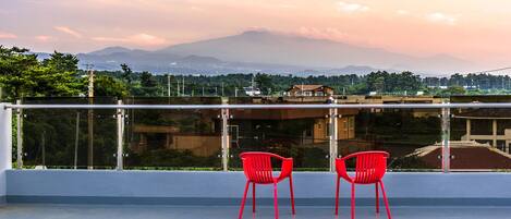 A balcony with two red chairs, a clear view of a mountain range, and a cityscape in the background.