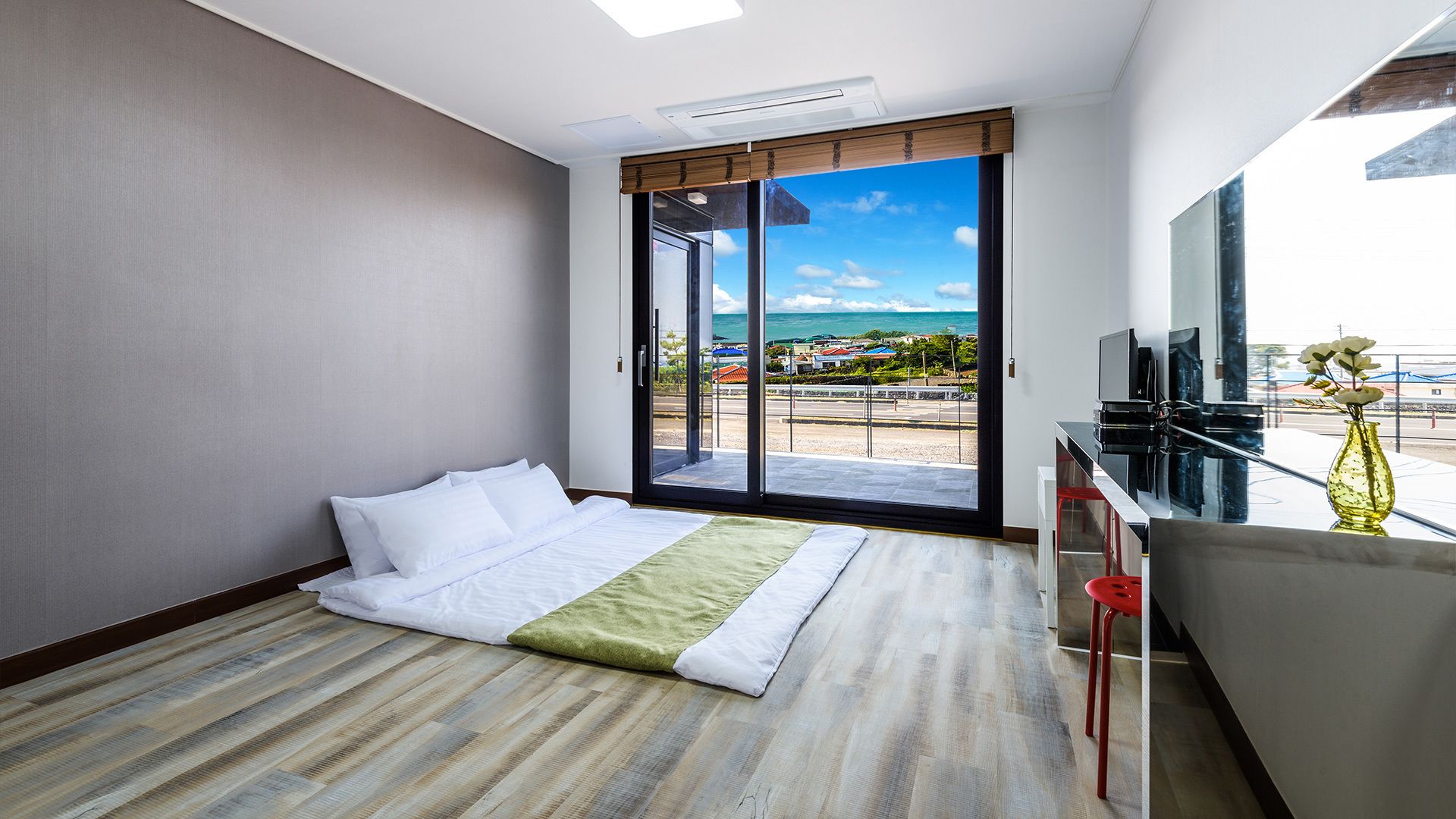 Habitación de hotel con cama, escritorio con televisor y una amplia puerta corrediza de vidrio que da a un balcón con vista a la playa y al océano.