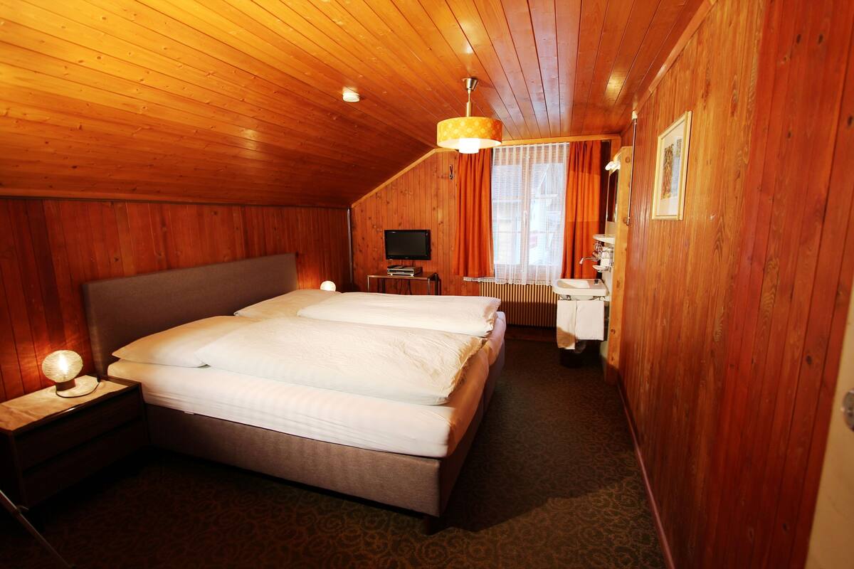 A room with a wooden ceiling, a bed with white bedding, a bedside table with a lamp, a television, and a bathroom with a sink and mirror.