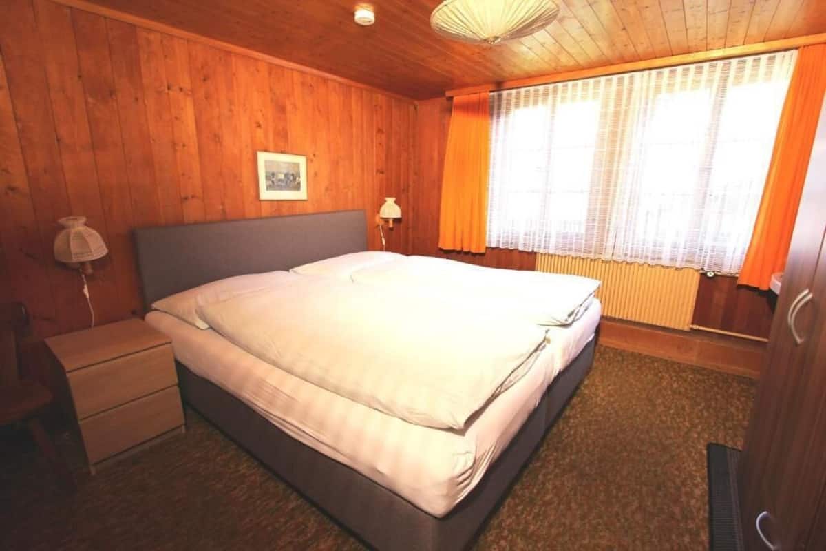 A wooden-paneled bedroom with a bed, a nightstand, a lamp, and a window with curtains.