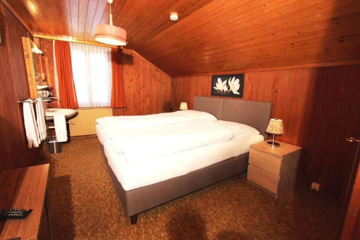 A wooden-paneled bedroom with a large bed, bedside tables, a radiator, and a window with curtains.