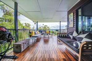 A covered outdoor deck with wooden flooring, a sofa, a dining table, and a grill.