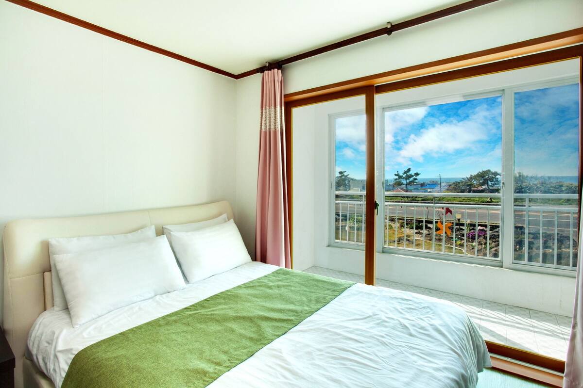 A bedroom with a large window, a bed with white linens, and a green throw blanket.