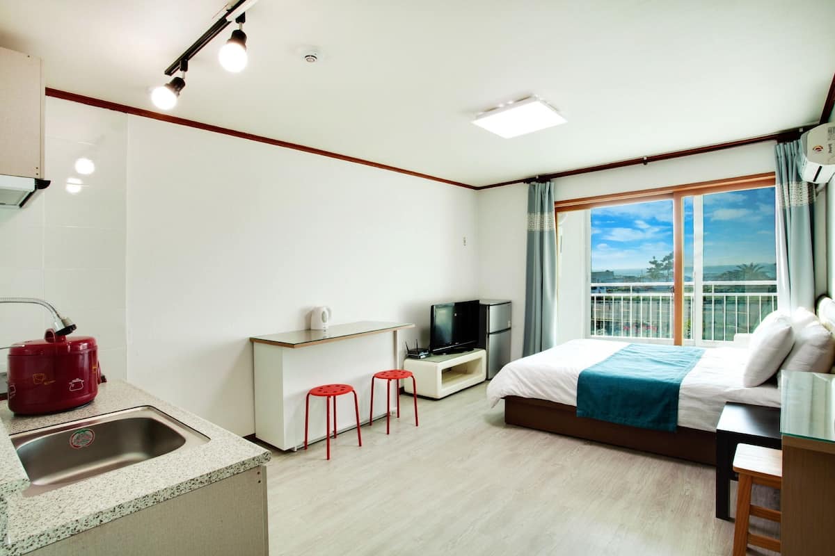 A hotel room with a kitchenette, a bed with a blue blanket, a TV, and a balcony with a view.