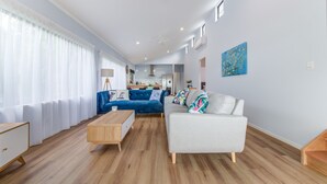 A spacious living room with a blue sofa, a white sofa, and a wooden coffee table. There is a kitchen in the background with a blue wall and a painting.