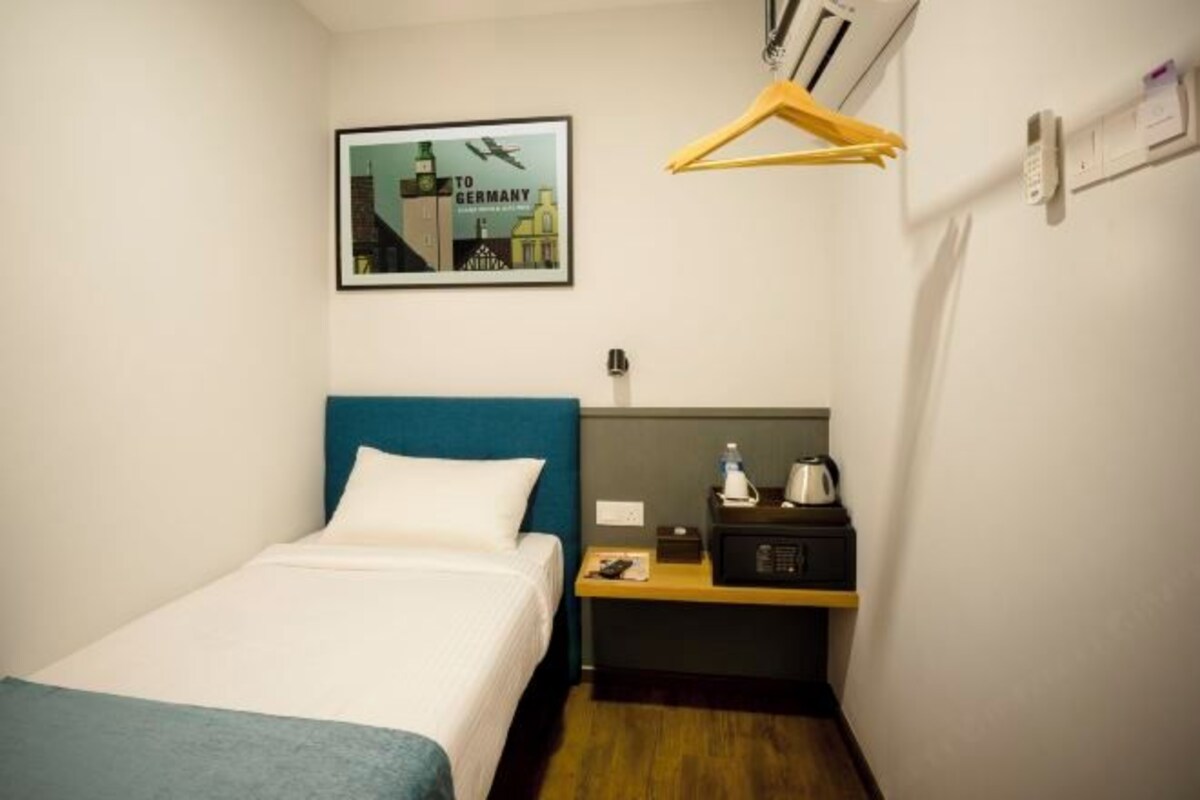 A small hotel room with a single bed, a bedside table with a kettle and a remote control, a framed picture on the wall, and a coat hanger.