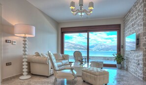 Ein Wohnzimmer mit einer Couch, Sesseln, einem Hocker und einem Couchtisch. Eine Lampe, ein Kronleuchter und ein an der Wand befestigtes Fernsehgerät sind vorhanden. Das Zimmer verfügt über ein großes Fenster mit Blick auf Berge und ein Gewässer.