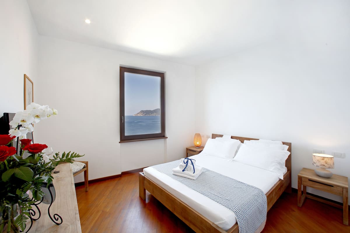 A bedroom with a wooden bed, white bedding, a bedside table with a lamp, a window with a view of the sea, and a vase with flowers.