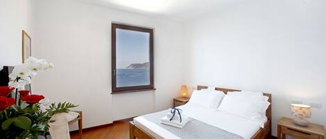 Un dormitorio con cama de madera, ropa de cama blanca, una mesita de noche con lámpara, una ventana con vista al mar y un jarrón con flores.