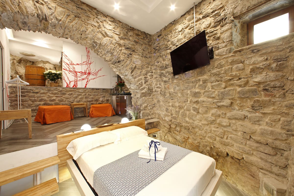 A bedroom with a stone wall, a bed, a wooden bench, a small table, and a TV mounted on the wall.