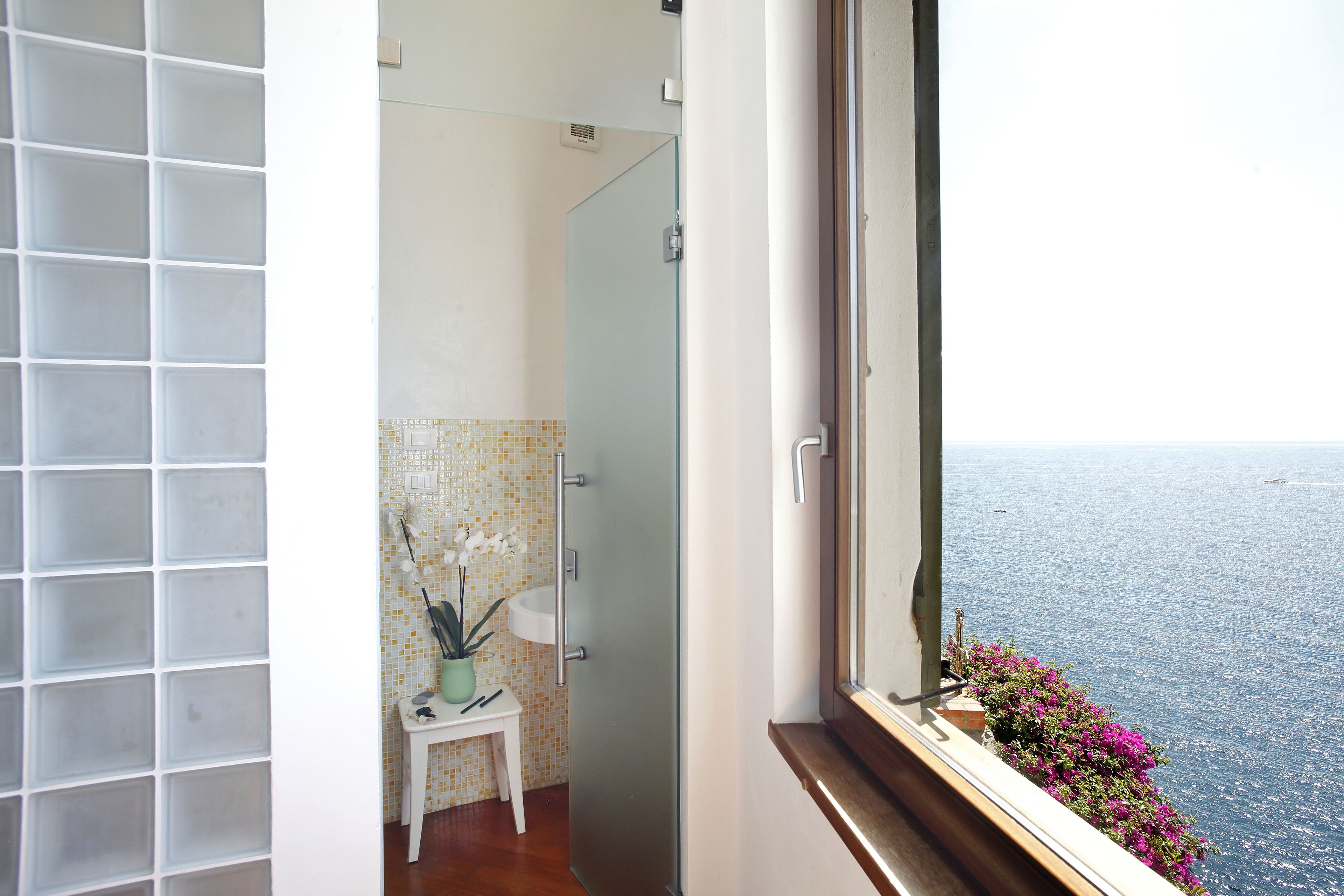 A bathroom with a white sink, a small table with a potted plant, and a view of the sea through a window.