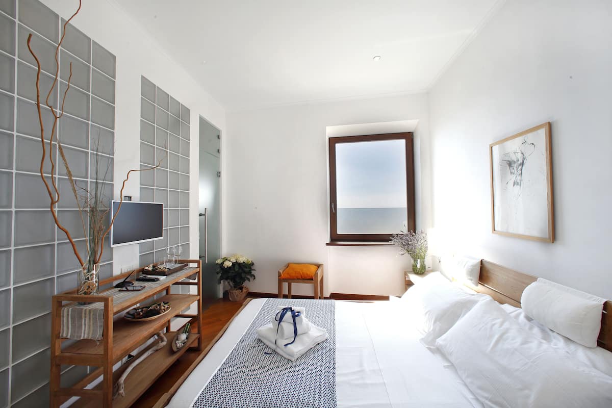 A modern hotel room with a large bed, a TV on a wooden stand, a small table with a vase of flowers, and a window with a view.