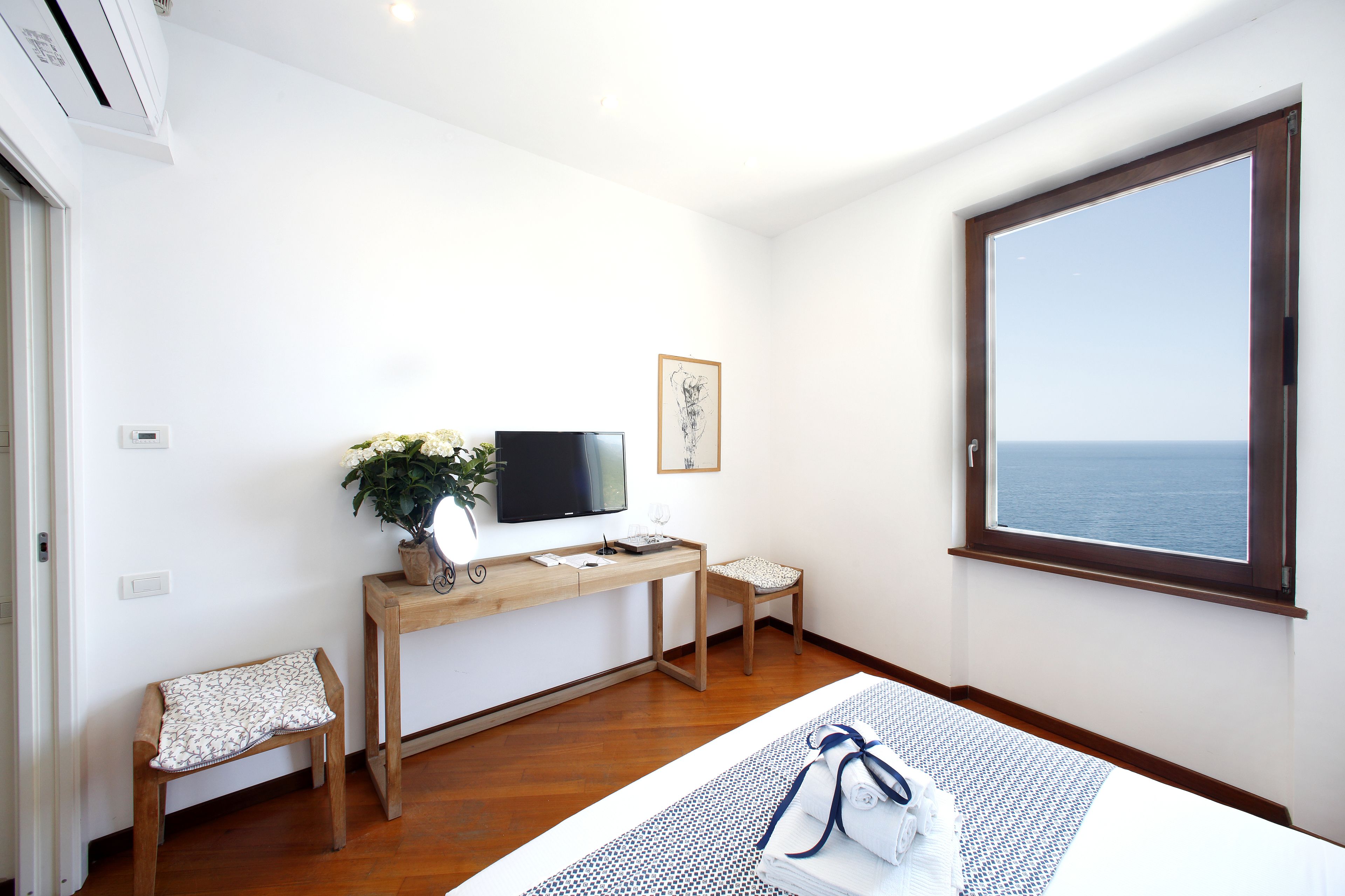 Habitación de hotel con un escritorio de madera, televisor, una pequeña silla, una mesita de noche con una toalla enrollada y un ventanal con vistas al mar.
