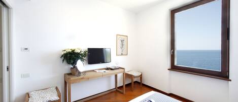 A hotel room with a wooden desk, a television, a small chair, a bedside table with a rolled towel, and a large window with a view of the sea.