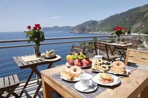 A table set with breakfast items and a vase of red flowers, overlooking a coastal view. - Arpaiu - Odeyo Sas (Riomaggiore)