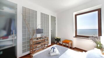 A hotel room with a bed, a desk with a lamp, a chair, a window with a view, and a plant.