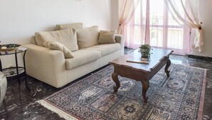 Uma sala de estar com sofá bege, mesa de centro de madeira, tapete estampado, quadro na parede e cortinas leves.