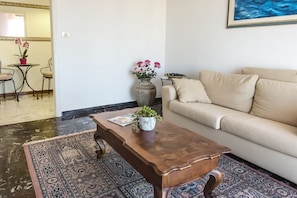Een woonkamer met een beige bank, een houten salontafel, een kamerkleed met een patroon, een kleine tafel met stoelen, en een vaas met bloemen.