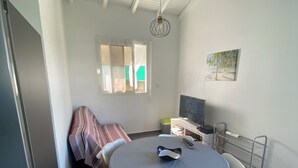 A dining area with a round table, chairs, a refrigerator, a window with curtains, a ceiling light, and a wall-mounted shelf with items.