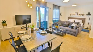 A modern living room with a sofa, coffee table, and dining area. There is a TV, a plant, and a window with curtains. - Split Luxury Collection (Split)