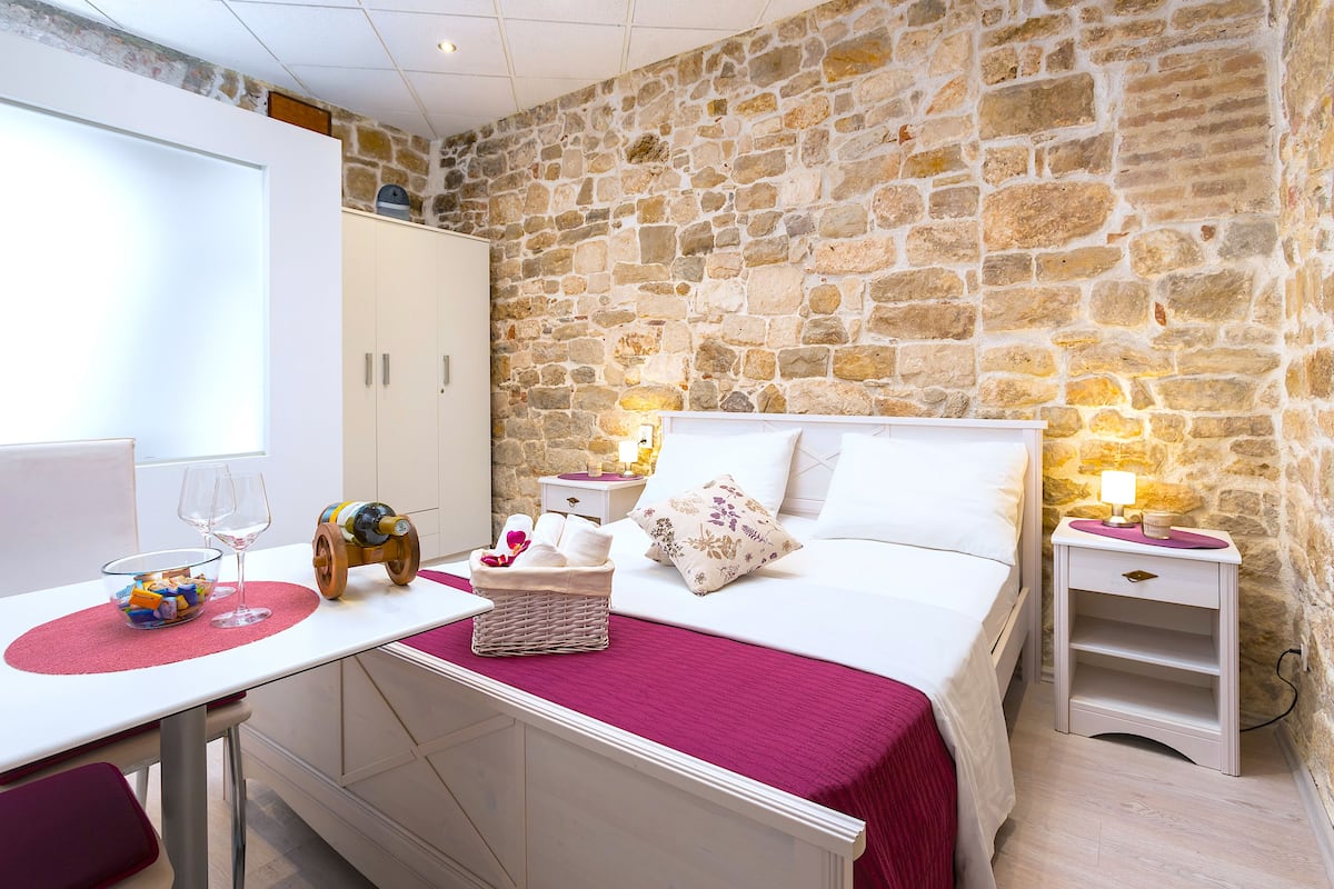 Un dormitorio con pared de piedra, una cama con manta roja, una mesita de noche con lámpara, y una pequeña mesa con cesta y copas de vino.