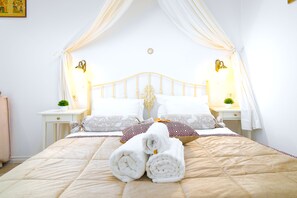 Una cama bien hecha con colcha color beige, almohadas blancas y una toalla blanca enrollada al frente. Hay dos mesitas de noche, cada una con una lámpara y una planta pequeña.