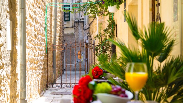 A narrow street with a table set with flowers and a glass of orange juice, a metal gate, and buildings with stone walls and greenery. - Gaius Aurelius Valerius (Split)