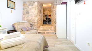 A bedroom with a stone wall, a bed with a beige cover, a white bedside table, and a TV mounted on the wall. - Gaius Aurelius Valerius (Split)