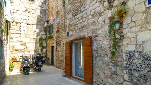 A narrow street with stone buildings, a parked scooter, and a shop with a wooden shutter. - Gaius Aurelius Valerius (Split)