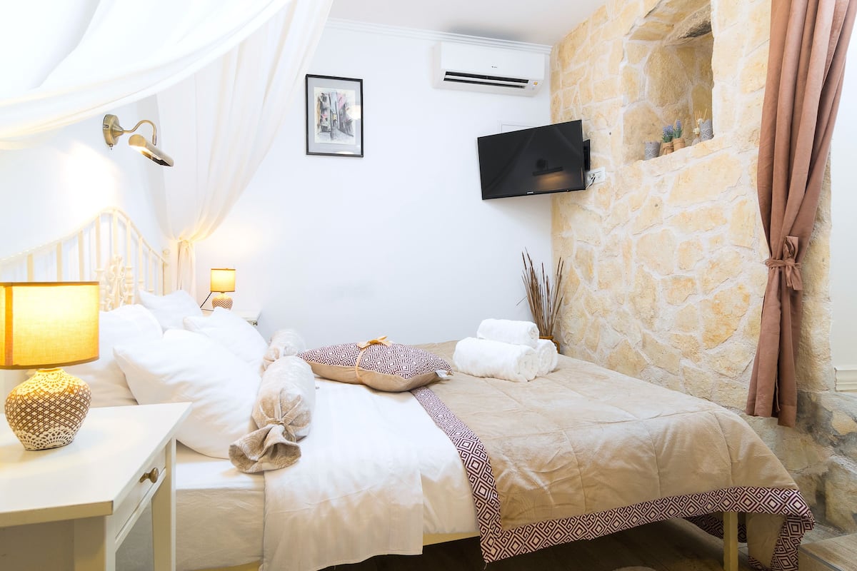 Una cama bien hecha con una colcha estampada, una lámpara de noche y un televisor montado en la pared.