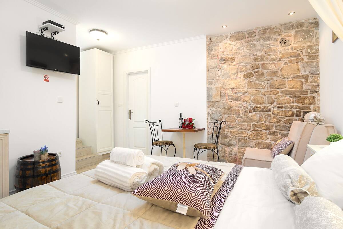 Un dormitorio con pared de piedra, un televisor de pantalla plana, una cama con almohadas estampadas y una pequeña mesa con sillas.