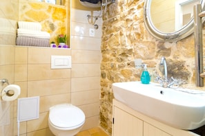 Un baño con pared de piedra, un lavabo blanco con dispensador de jabón, un inodoro y un perchero con toallas dobladas.