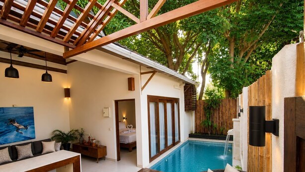 Un área de piscina moderna al aire libre con terraza de madera, una silla de lounge de mimbre y vista a un dormitorio con cama y baño.