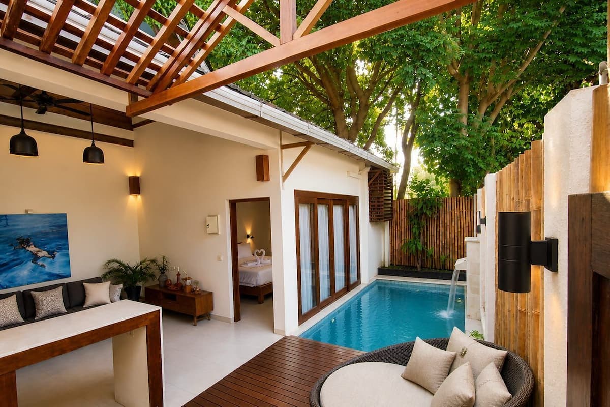 A modern outdoor pool area with a wooden deck, a wicker lounge chair, and a view of a bedroom with a bed and a bathroom.