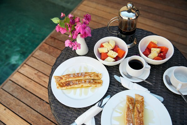 A table with breakfast items including crepes, fruit, coffee, and a flower vase by a pool.