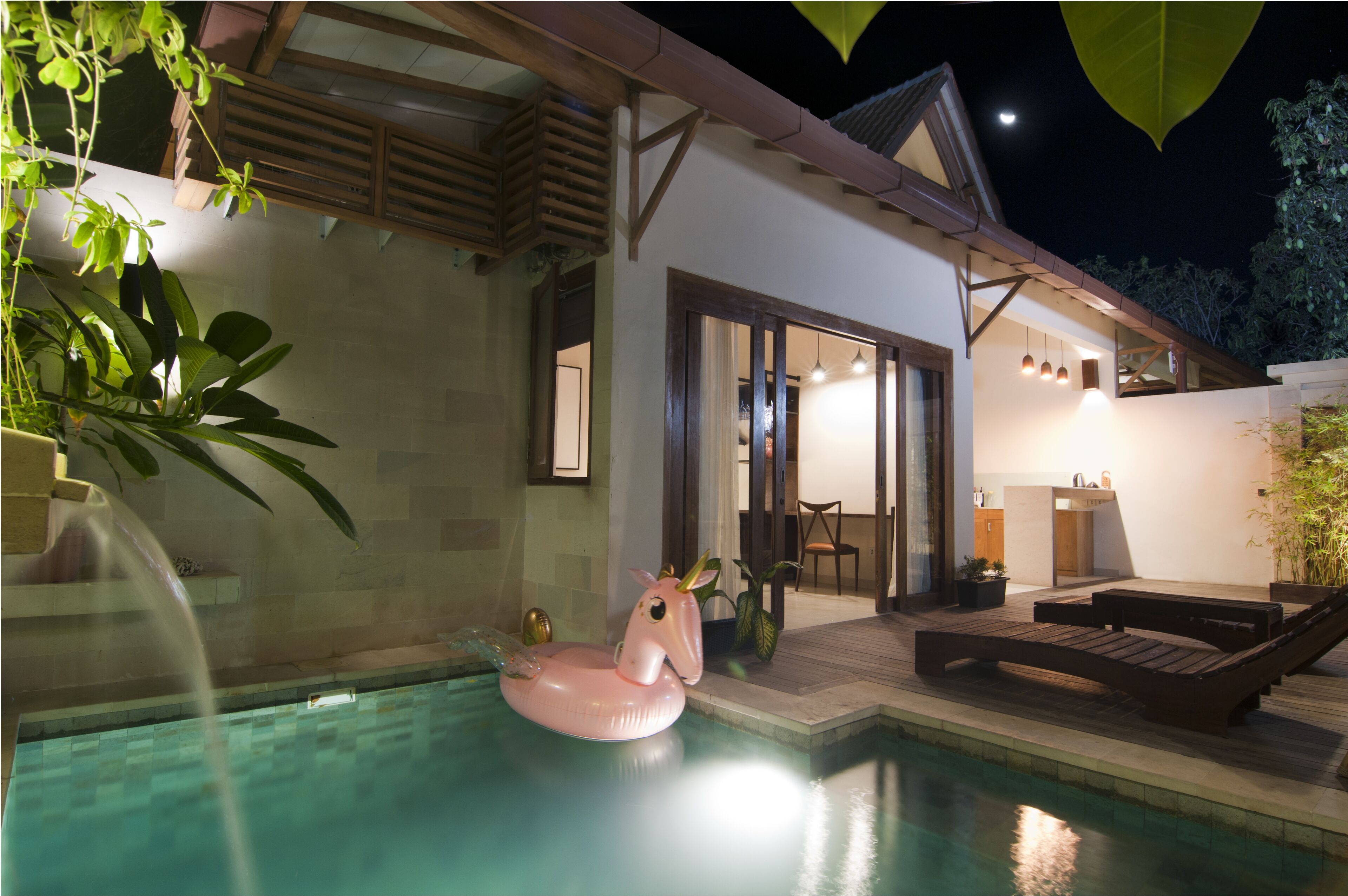 Une piscine agrémentée d’un flotteur en forme de flamant rose, un espace de détente en bois avec des sièges, et une maison moderne avec une cuisine ouverte.