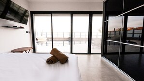 A modern hotel room with a large bed, a flat-screen TV mounted on the wall, a small table, and a large sliding glass door leading to a balcony with a view of the city. - Villa Daniela (Eilat)