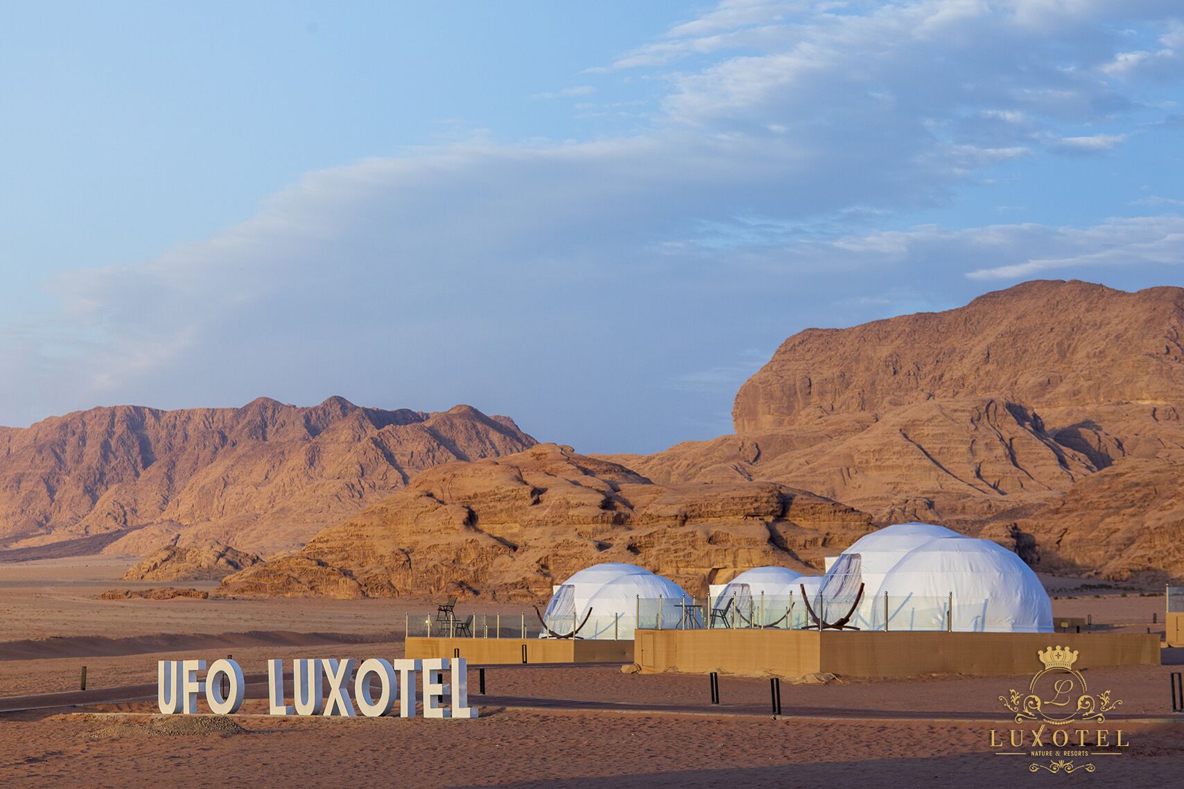 A desert landscape with a hotel named 'UFO LUXOTEL' featuring large white domes, a clear sky, and mountains in the background.