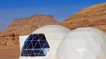 Una estructura en forma de cúpula blanca con un diseño de ventanas geométricas, situada en un paisaje desértico con colinas rocosas.