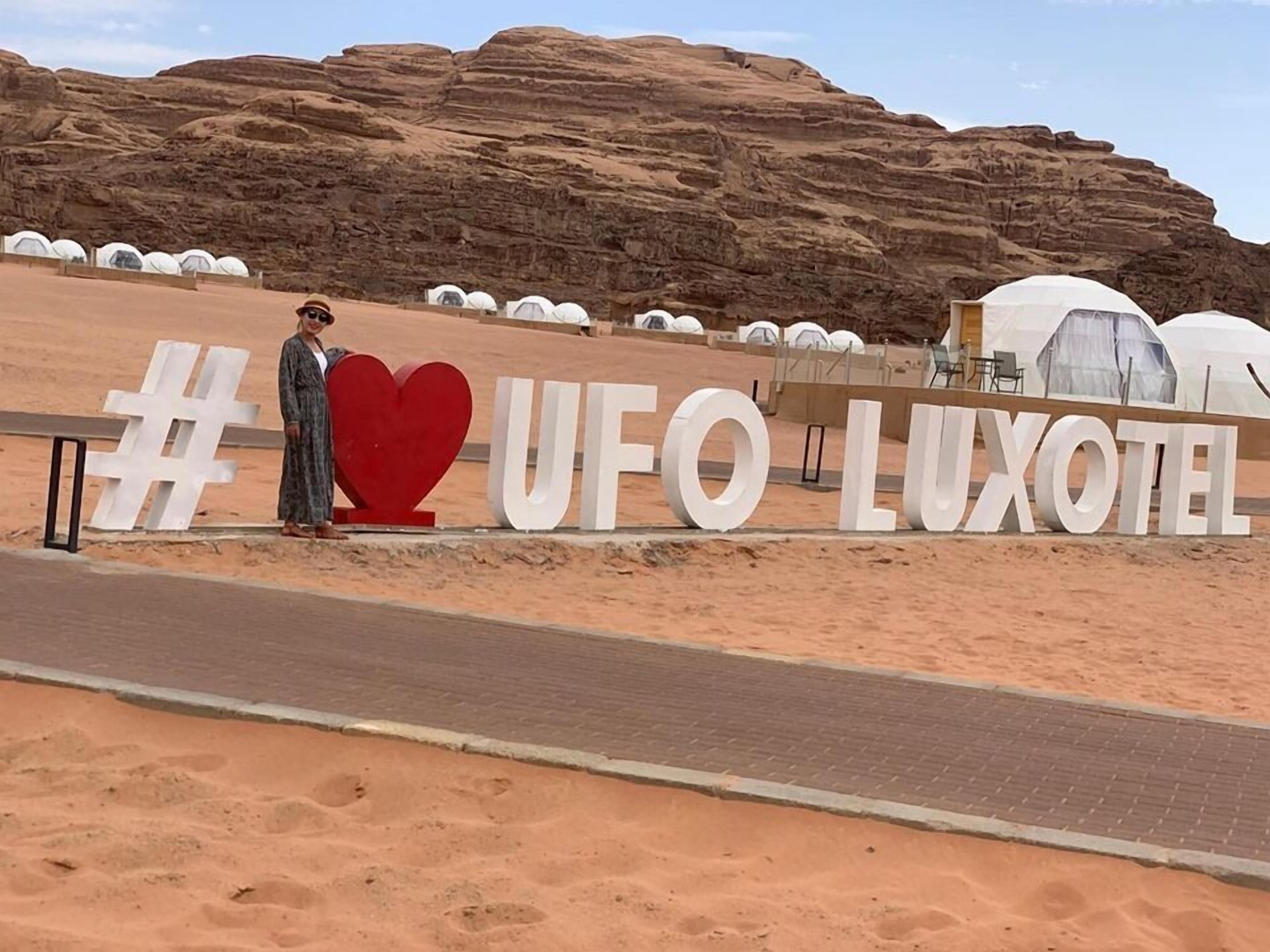 Paisaje desértico con tiendas de campaña identificadas como "UFO LUXOTEL" y una persona de pie frente a un letrero grande con forma de corazón.