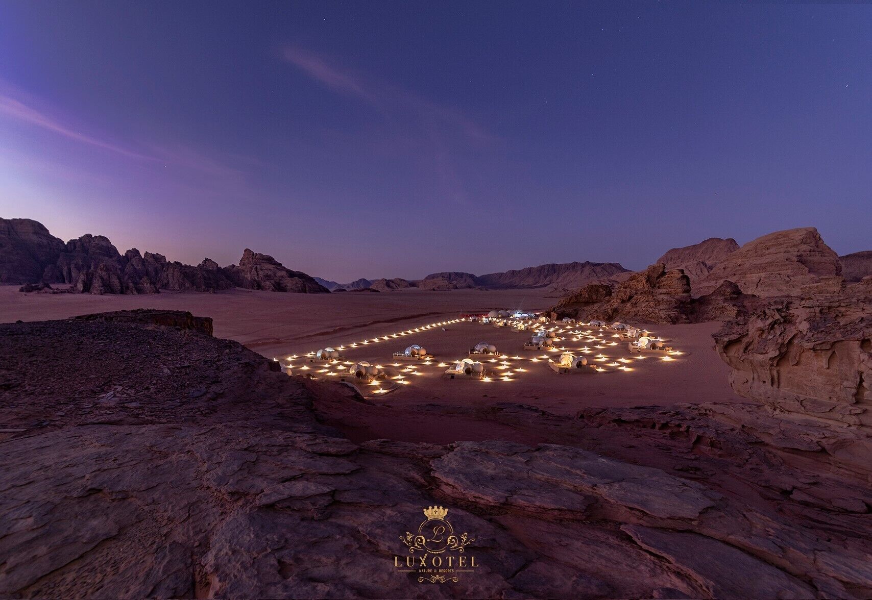 A desert landscape with a heart-shaped arrangement of illuminated tents, suggesting a luxury hotel setting.